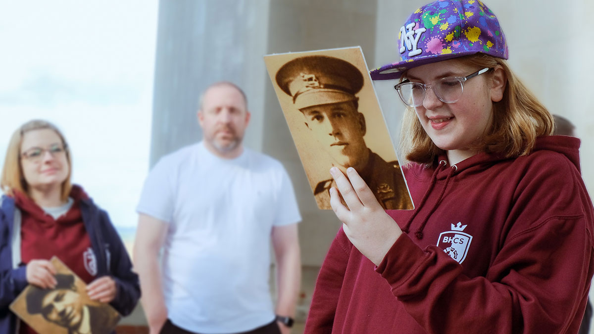 Students from Brighton Hill Community School reads aloud a soldiers story whilst at a World War memorial. 2 adults stand and look on in the background.