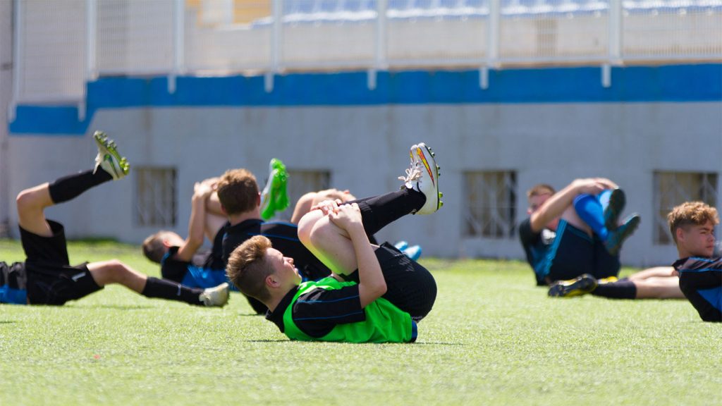 A boys football team stretches whilst on the green pitch on a sunny day.