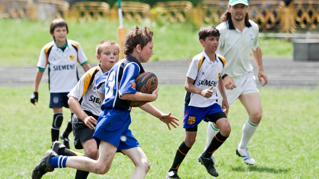 A rugby player in the foreground runs past 3 opposing rugby players in the background on a green pitch on a sunny day