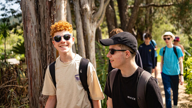 Two male students walking in a forest while talking, one of which is smiling the camera