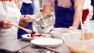 Close Up Of Woman Putting Topping On Pancake In Cookery Class As Adult Students Look On