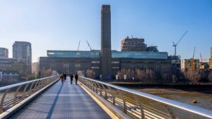 Visiting Tate Modern in London on a bright day. People stroll across a bridge towards Tate Modern, a well-known gallery in London. The sun shines brightly, highlighting the modern architecture of the building and surroundings.