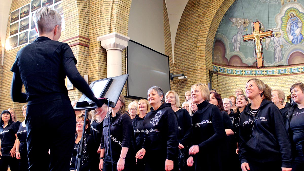 A choir, all dressed in black with a white logo on their clothing, practise with their choral director in a church