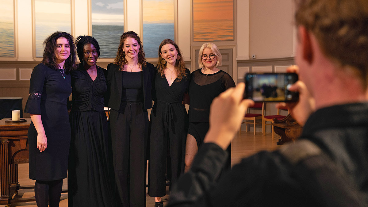 Five ensembles members, all in black, pose with linked arms around each others back to a photo being taken on a phone in the foreground