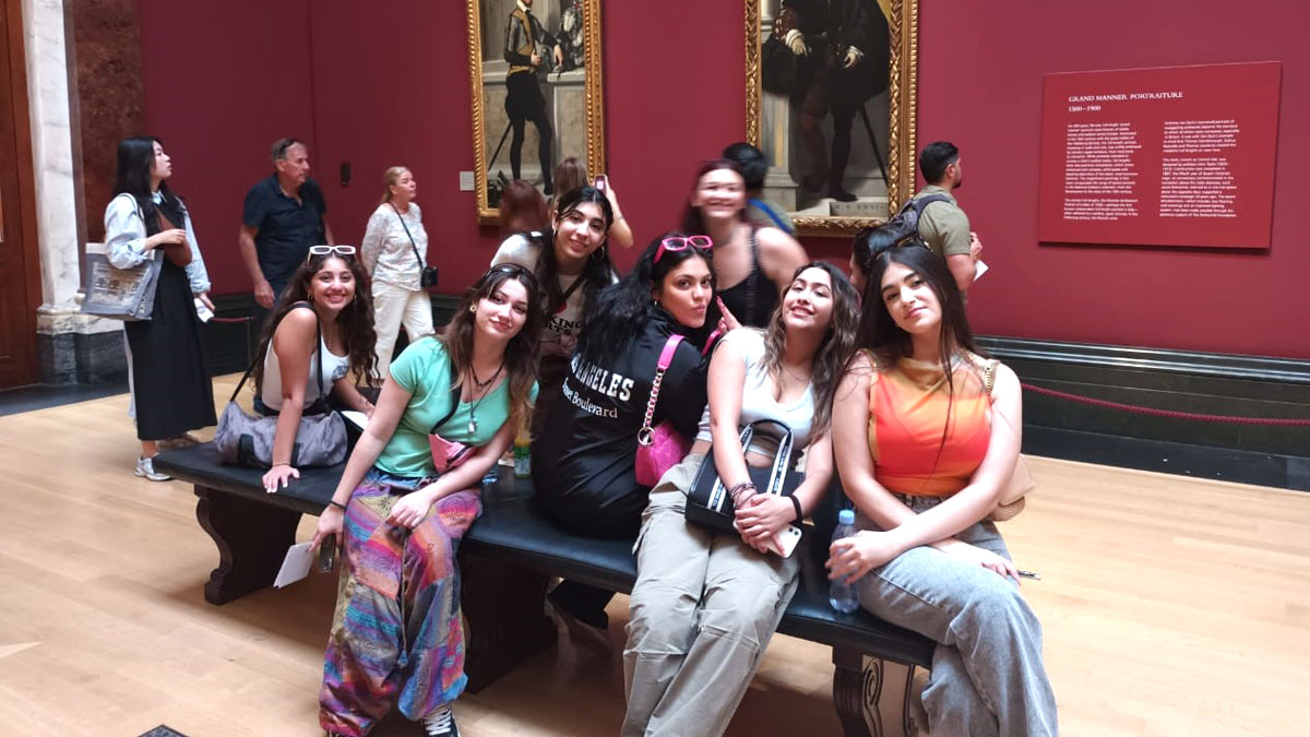 Girl students pose for a photo, whilst sitting down on a central bench in a red room with paintings, in a museum in London.