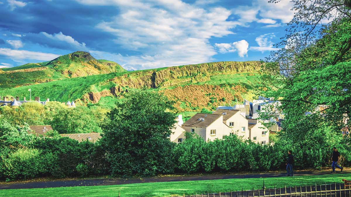 Image of green landscape, with building in the mid-ground and a blue, partially cloudy sky, of Holyrood Park