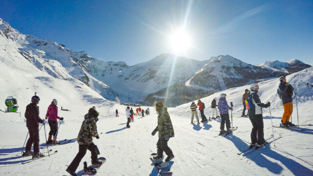 Skiers and snowboarders stand around and chat, with a high sun, mountains and white slopes