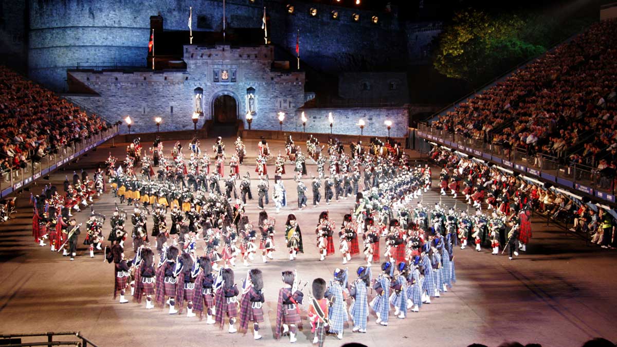 A military band with bagpipers, drums, banner holders, performs in formation in a night time Edinburgh Tattoo with Edinburgh Castle in the background.