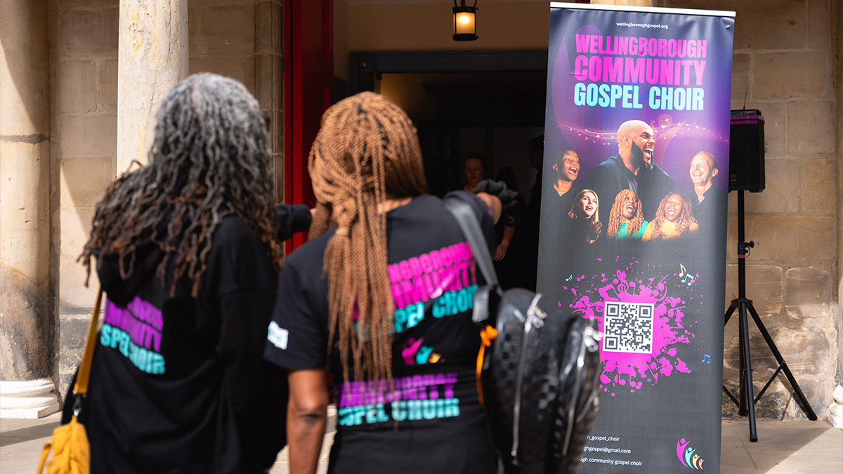 Wellingborough Community Gospel Choir outside of Canongate Kirk, pointing towards their promotional banner during the Fringe Festival