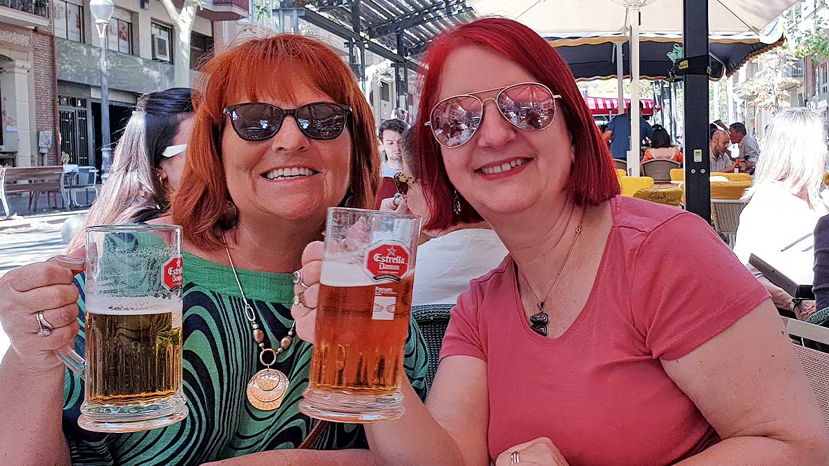 2 choir members pose with sunglasses on and beers in hand in a bar on a street in Barcelona.