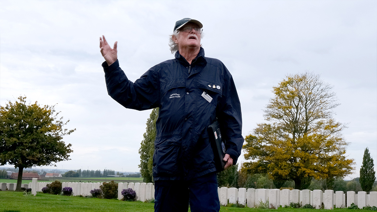 Trevor Booker, History Tour Guide, talks to a history tour off camera, whilst a World War graveyard is in the background.