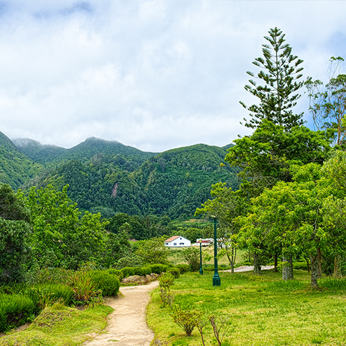 A pathway leads to green trees, with a building in the midground and high, green covered mountains in the background, of Furnas Valley.