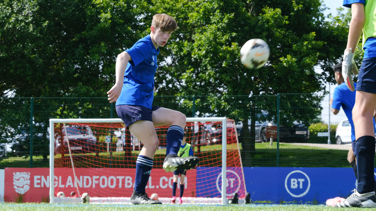 Player kicking a football on a pitch with goal net in the background