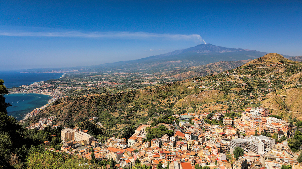 Mount Etna smokes in the background with green fields and houses unfolding beneath it.