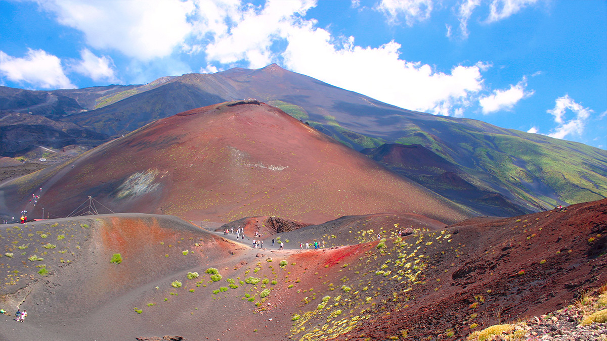 Mount Etna landscape with trekkers in the midground and a blue, cloudy sky.