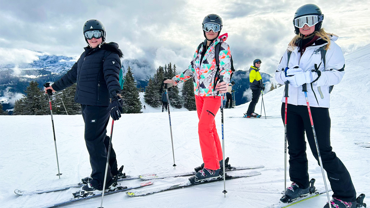 Three students pose and smile in their ski gear, ski poles, and mountains behind them