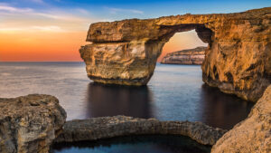 A scenic view of the Azure Window, a natural arch in Malta. The sun is setting, casting a warm glow on the rock formation and the surrounding ocean.