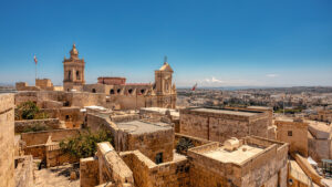 A panoramic view of the Citadel of Victoria in Gozo, Malta. The fortified city is perched on a hilltop, overlooking the surrounding countryside and the sea. The prominent St. George's Basilica dominates the scene.