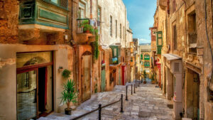 A narrow, winding street in Valletta, Malta, with colourful buildings and balconies. The street slopes downwards, with stairs leading to the bottom.