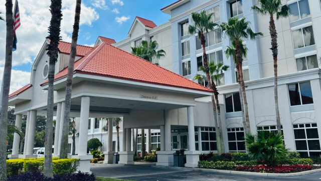 The exterior of a hotel with a red tiled roof, white columns, and blue accents. Palm trees line the driveway, and an American flag is flying.