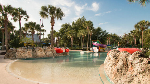 A hotel pool surrounded by palm trees. There are rock formations with water features and a colorful water slide in the background.