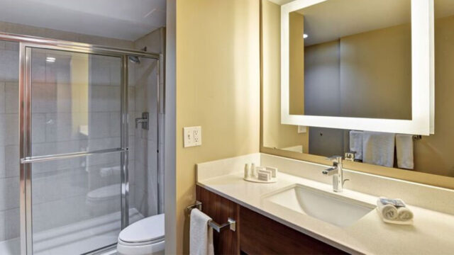 A hotel bathroom with a glass-enclosed shower, a sink with a countertop, a mirror with lights, and a toilet. Towels and toiletries are provided.