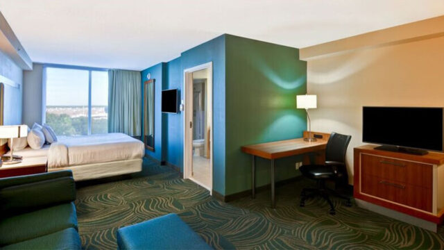 A hotel suite with a king bed, a sofa, a desk, a television, and a window with curtains. The walls are painted teal, and there is a door leading to a bathroom.
