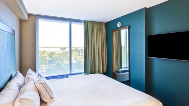 A hotel room with a king bed, a television, a large window with curtains, and a mirror on the wall. The wall behind the bed is painted teal.