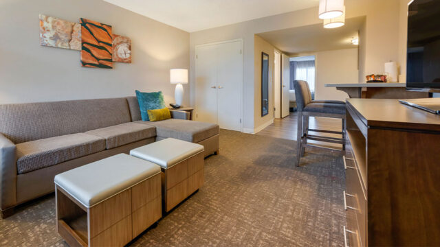A hotel suite living area with a sectional sofa, two ottomans, a coffee table, and a lamp. There is a wall-mounted television and a doorway leading to a bedroom and kitchenette area.