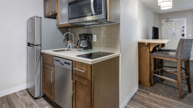 A hotel kitchenette with stainless steel appliances, including a refrigerator, microwave, coffee maker, and dishwasher. There is a countertop with a sink and electric stovetop, and a bar stool sits at a small counter.