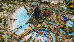 An aerial view of a large water park with a towering volcano at the center. The volcano features multiple water slides and attractions, surrounded by pools, wave pools, and lounging areas.