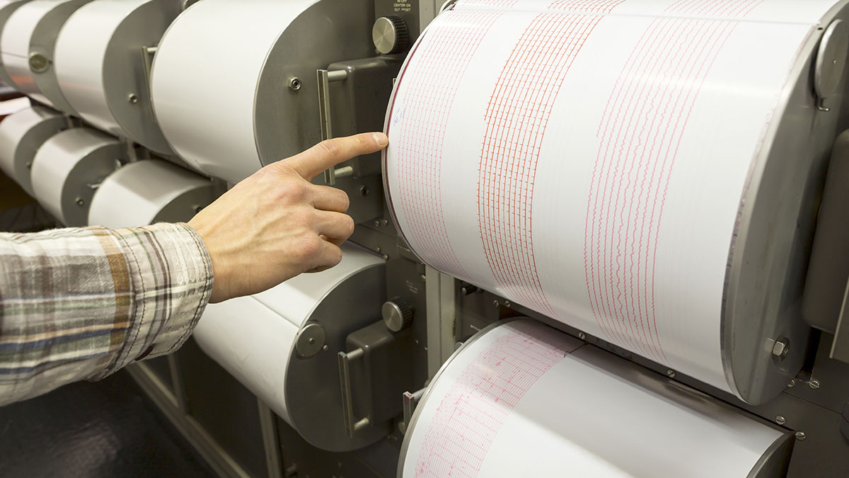 Seismograph recording earthquake human hand. Seismograph records an earthquake on the sheet of measuring paper. Seismological device for measuring earthquakes. Earthquake wave on graph paper. Human finger showing a detail of the earthquake.