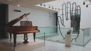 A museum exhibit featuring an ornate wooden grand piano displayed on a raised platform. Behind it, a white wall shows a large black arrow pointing right. To the right side of the space, a glass railing surrounds an opening to a lower level, and a tall wall features an oversized black outline of a hand filled with handwritten text. A small interactive display stands in front of the hand illustration. Track lighting illuminates the modern, minimal gallery interior.